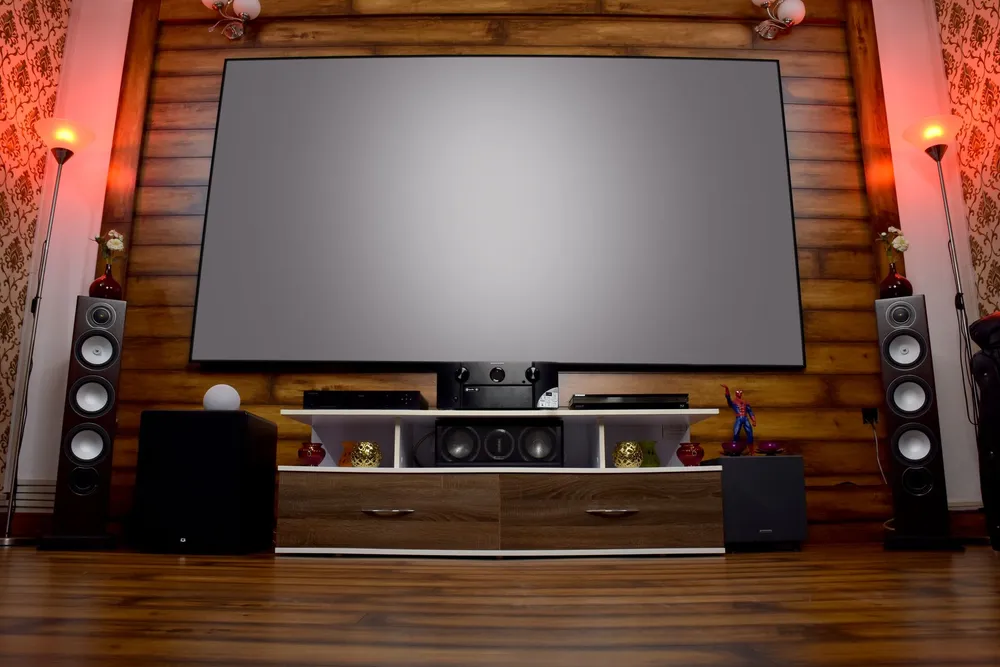 Salon home cinéma avec un grand écran de projection, une enceinte colonne de chaque côté, un caisson de basses posé au sol et un mur habillé de bois